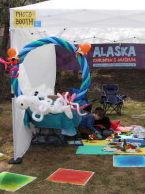 A white pop up tent outdoors with Alaska Children's Museum banner and colorful toys. 