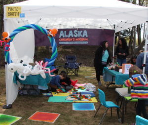 A white pop up tent outdoors with Alaska Children's Museum banner and colorful toys.