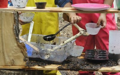 Two children play in a mud kitchen made of split logs. The child on the left wears yellow, rubber coveralls. The child on the right wears pink and has two brown braids.