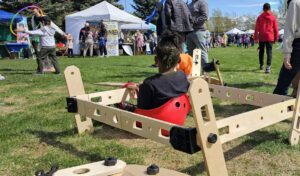 Young African-American child plays at an outdoor event in a car made of wooden building panels. Their hair is in short locs held up in a ponytail and shaved on the sides. People are walking by and another child plays with hoop toys in the background