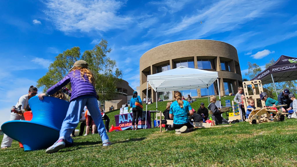 Children playing outdoors with spinning top chairs and other museum toys in front of the Loussac Library in Anchorage