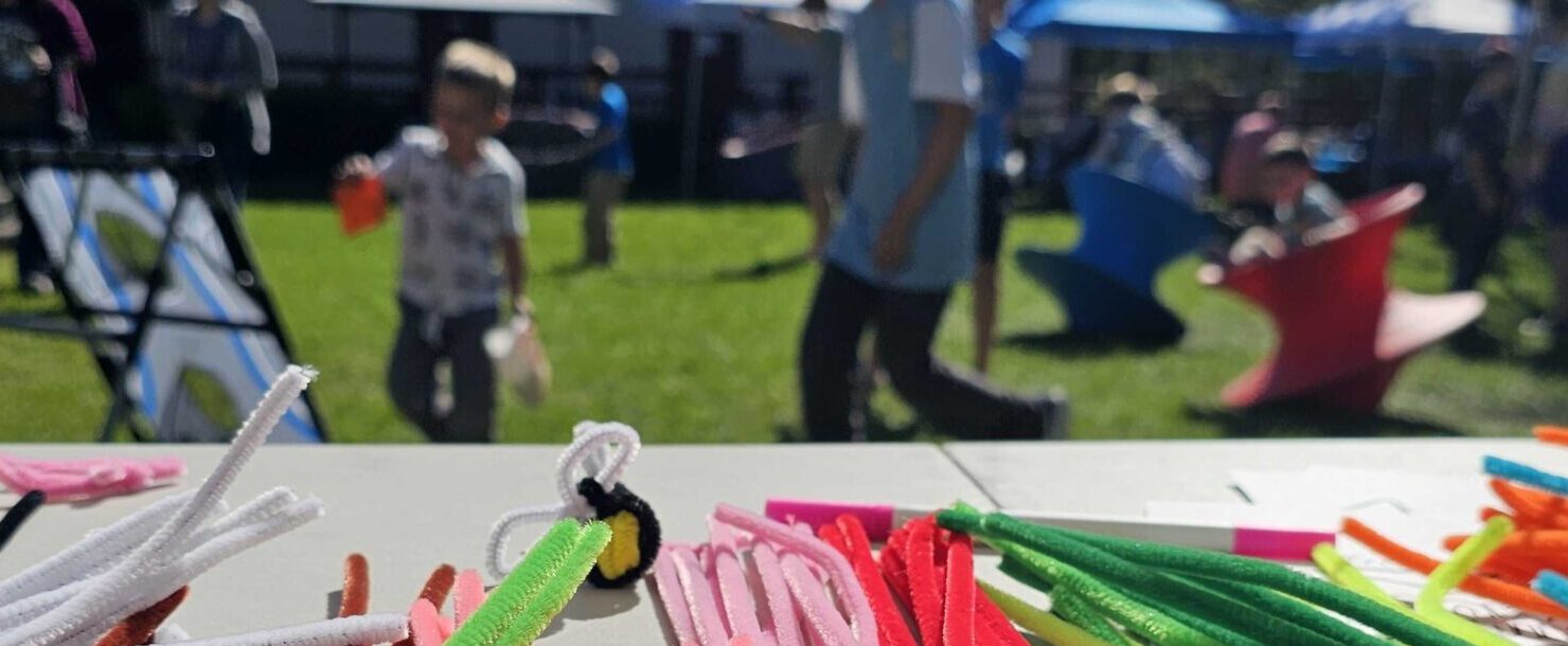 Colorful row of craft pipe cleaners in the foreground. In the background and out of focus, kids play outdoors with a beanbag toss and spinning chairs.